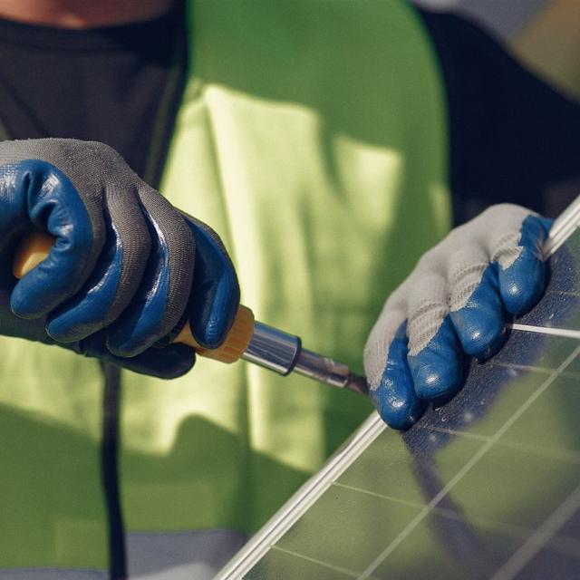 Eine Person in einer Warnweste inspiziert ein Solarmodul mit blauen Arbeitshandschuhen.